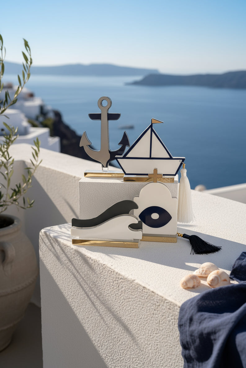 Σύγχρονα διακοσμητικά γούρια από πλεξιγκλάς σε κυκλαδίτικο φόντο: άγκυρα από χρυσό καθρέφτη, καραβάκι με navy περίγραμμα και φούντα, κύμα σε λευκό-γκρι, και ναός με ματ μάτι – όλα σε διάφανη και χρυσή βάση.