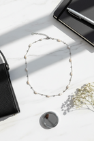 Necklace on a white surface with a black handbag, notebook, pen, and small round mirror.