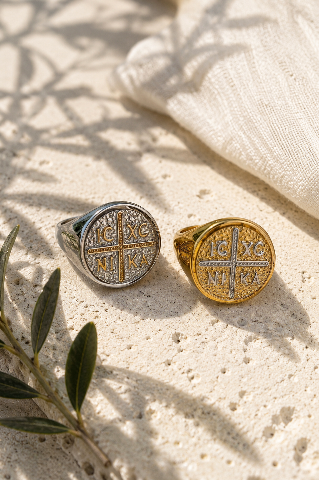 Two round signet rings with religious symbols, one silver-toned and one gold-toned, displayed on a textured beige stone surface with soft leaf shadows and a folded cream fabric in the background.