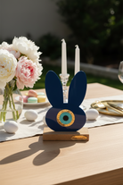 Decorative blue bunny figurine with a gold eye on a wooden base, placed on a table with flowers and candles.