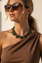 Woman wearing a brown dress and green heart-shaped necklace with sunglasses and gold earrings.