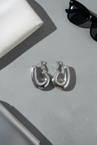 Silver hoop earrings on a light gray surface with sunglasses and a book in the background.