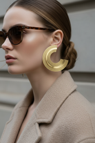 Woman wearing gold plexiglass earrings and sunglasses with a neutral background