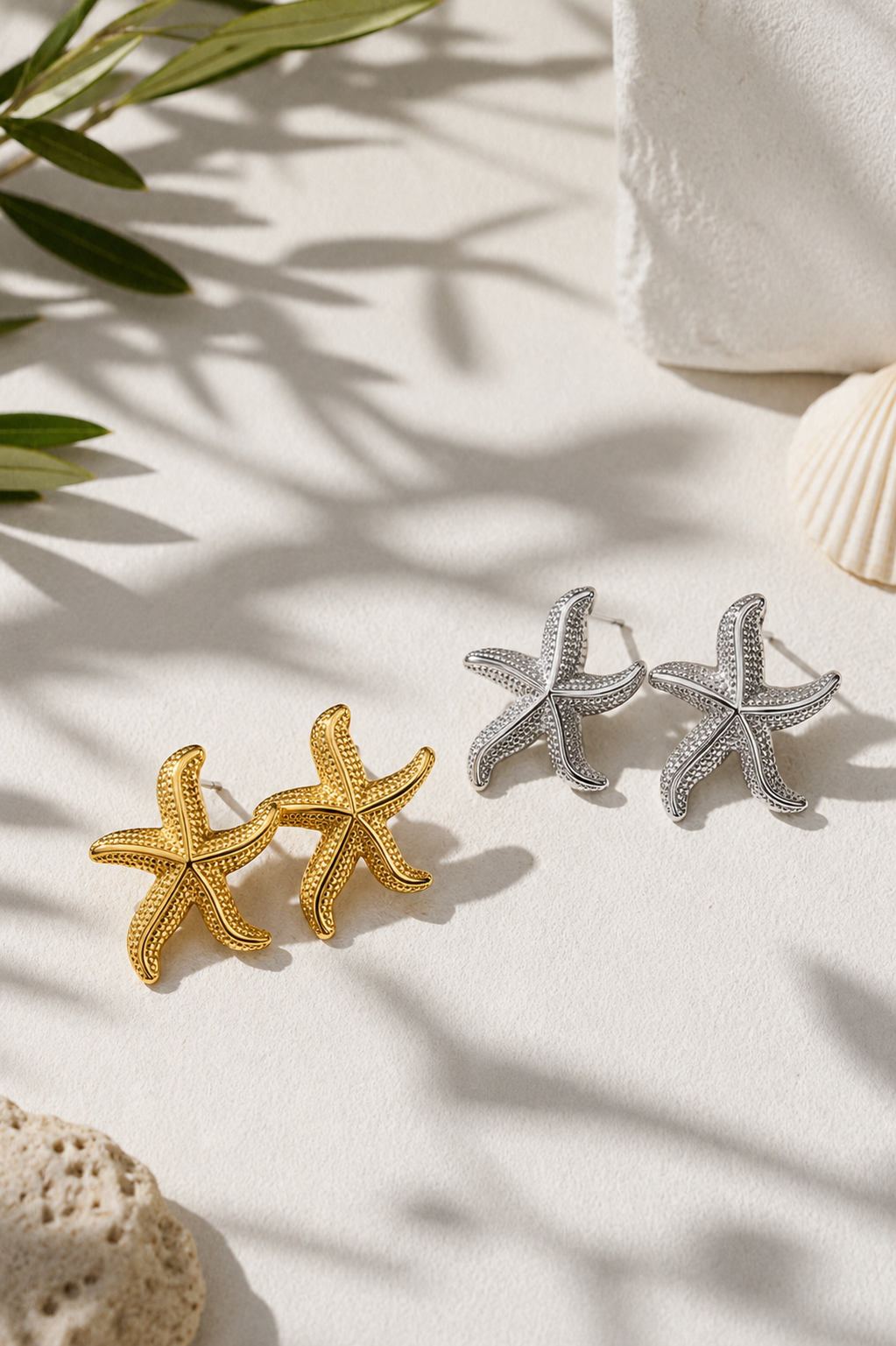 Two starfish-shaped stud earrings, one in yellow gold and one in silver, displayed on a light textured surface with soft shadows, decorative leaves, and shell props.