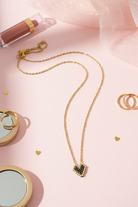 Gold stainless steel necklace with a heart miyuki pendant on a pink surface with lip gloss and rings.