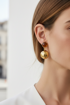 Close-up of a person wearing gold and amber round earrings against a blurred background