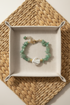 Green beaded bracelet with a pearl on a beige tray, placed on a woven mat.