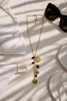 Élira Coin Y Necklace μαύρο χρυσό σε καλοκαιρινό flatlay με bikini και γυαλιά ηλίου