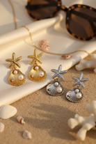 Pair of starfish and shell stainless steel earrings on a sandy surface with shells and sunglasses in the background, So Cute by DImi