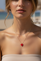 Close-up of a woman wearing a gold necklace with a red heart miyuki pendant on a beach.
