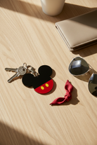 Keychain with Mickey Mouse design on a wooden surface with sunglasses and wallet.