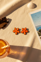Flower-shaped earrings on a sandy surface with a drink and card in the background