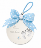 A personalized baby keepsake ornament featuring a round silver-toned plate with cutout baby footprints and a customizable text field, adorned with a light blue and white star-patterned ribbon bow and a blue accent stone, on a white background.