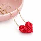 Red beaded heart pendant on a gold chain with a pink dish in the background.