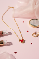 Gold stainless steel necklace with a red miyuki heart pendant on a pink surface with lip products and heart-shaped confetti.