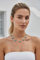 A woman with blonde hair in a ponytail wearing a silver chain necklace with turquoise starfish charms, paired with a white strapless top against a bright, softly blurred background.