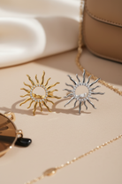 Sun-shaped earrings on a beige surface with sunglasses and a handbag in the background