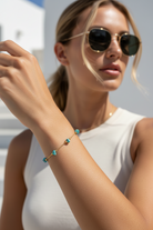 A woman wearing round sunglasses and a white sleeveless top wears a delicate gold bracelet on her wrist. The bracelet features small, faceted turquoise-colored beads set in gold.