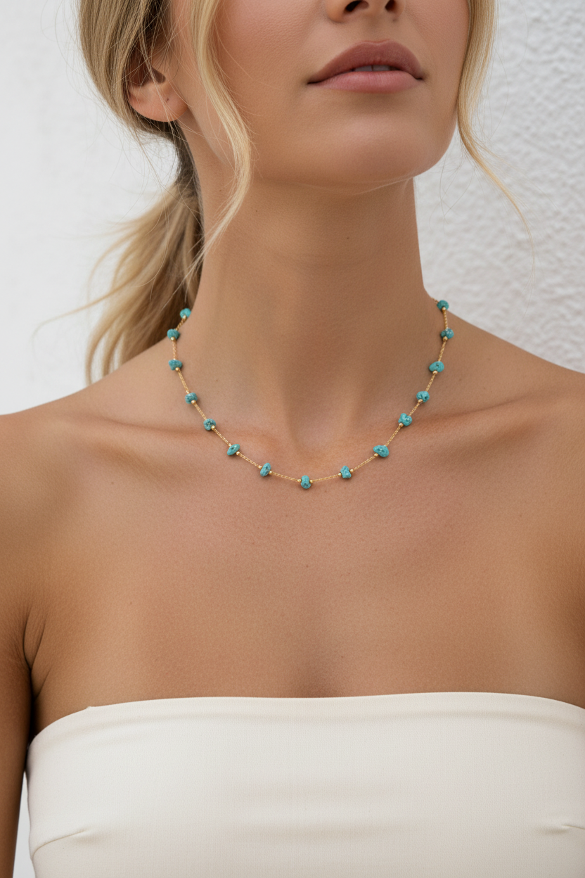 A close-up of a model wearing a delicate gold chain necklace adorned with small turquoise blue beads, paired with a white strapless top against a textured white wall.