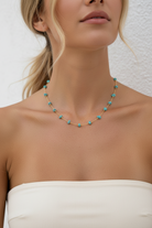 A close-up of a model wearing a delicate gold chain necklace adorned with small turquoise blue beads, paired with a white strapless top against a textured white wall.
