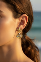 Close-up of a woman's ear wearing a gold earring with a starfish and shell design, against a blurred beach background, So Cute by Dimi